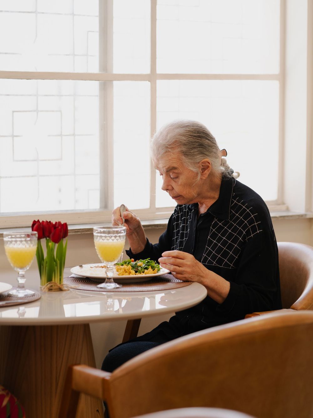 An older woman enjoying a plated meal with juice by a bright window